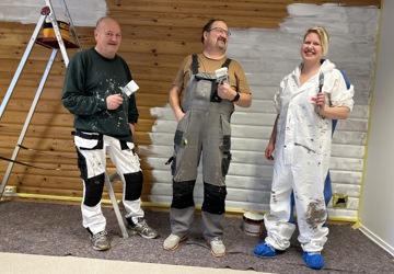 To menn og en kvinne i arbeidstøy som står foran en vegg som er malt halveis i hvitt. De tre personene holder opp hver sin malekost. Munter stemning. 